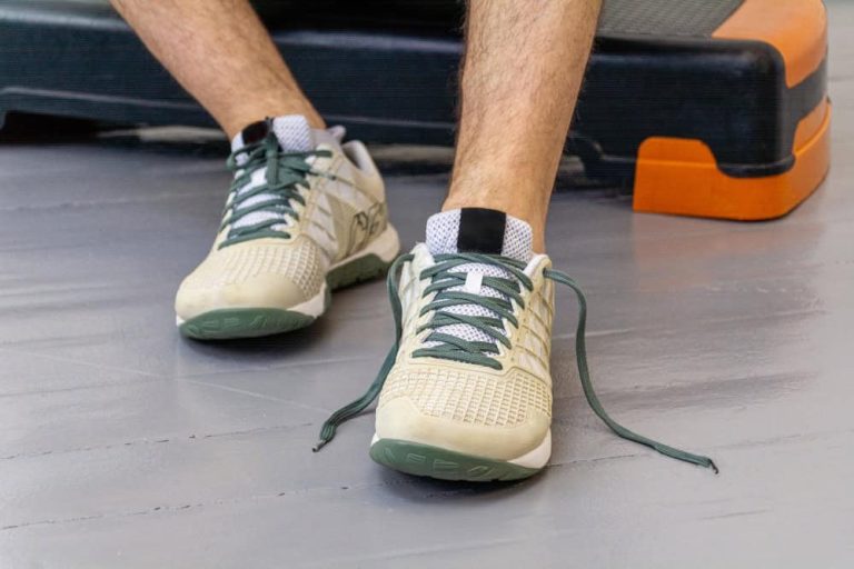 Primer plano detallado de las piernas de un hombre sentado en el suelo del gimnasio, mostrando un par de zapatillas de calzado barefoot para crossfit de color crema y beige. Una zapatilla tiene los cordones verde oliva atados y la otra los tiene desatados. Se aprecian la suela minimalista verde oliva y el fondo con una plataforma de salto naranja y un suelo de hormigón gris, destacando el diseño del calzado.